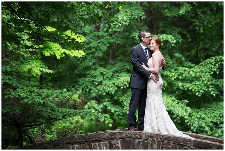 Bride and Groom stand on bridge surrounded by greenery embracing each other