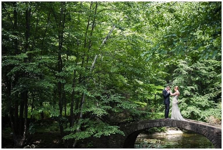 Bride and Groom stand on bridge over small pond while he kisses her hands