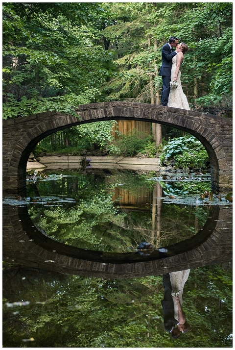 Bride and groom stand on small bridge while kissing, and the bridge is symmetrically reflected in the pond for the wedding anniversary photo session