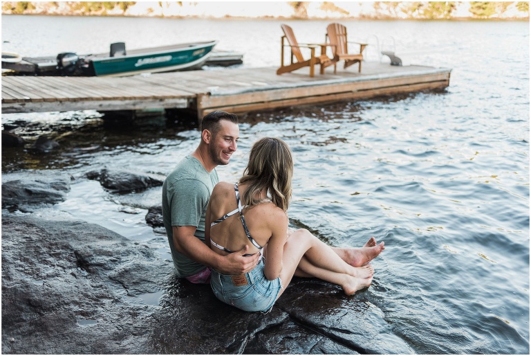 Man and woman sit on rocks on the shore of their cottage engagement session. Dock and yellow muskoka chairs are in the background