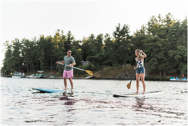 Man splashing woman with his paddle while the couple paddle boards across the lake for their engagement session