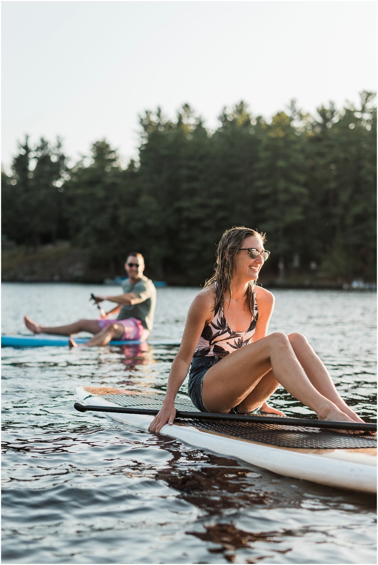 Woman sitting on paddle board while boyfriend is in the background smiling on his paddle board during their cottage engagement session