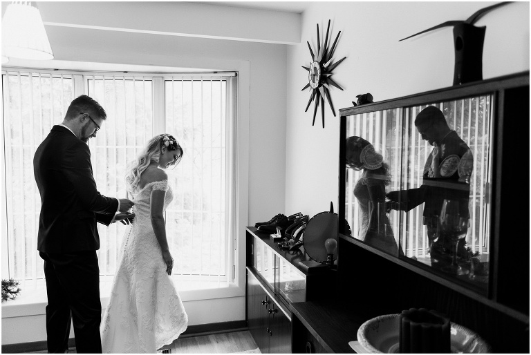Bride and groom are seen in reflection of the cabinet in their living room as groom does up bride's wedding dress
