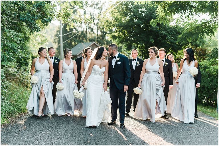 Bride and Groom kissing each other with their wedding party walking down a backlit street all together outside of the Ancaster Mill wedding