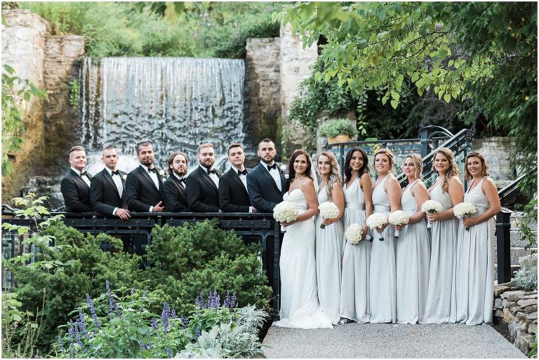 Bride and groom pose with their wedding party on front of waterfall at The Ancaster Mill