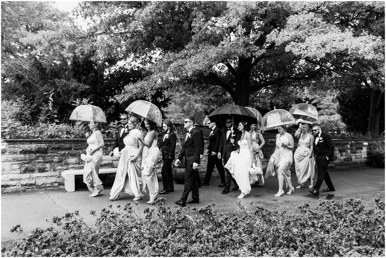 Black and white photo of wedding party walking with umbrellas during wedding photos at Hendrie Park inside of Royal Botanical Gardens