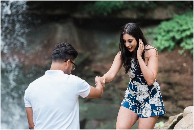 Guy helping his fiancée walk across some rocks in front of Hamilton waterfall 