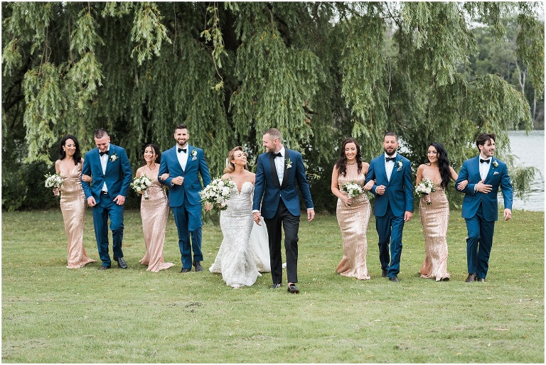 Wedding party walks on grass in their rose gold sequin dresses and navy suite with black bowties