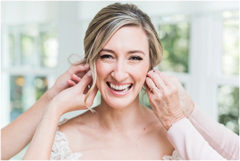 Bride has assistance putting on both of her earrings as she smiles brightly