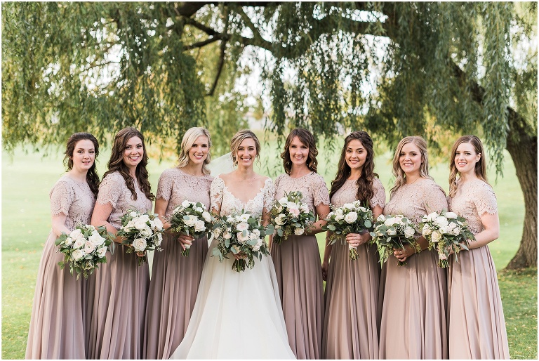Bride and her bridesmaids who are wearing soft pink dresses are all looking at the camera outside of the wedding venue