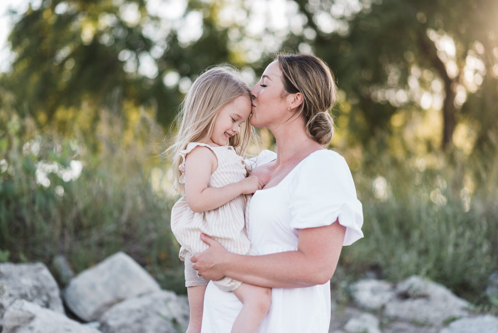 Mother holding her daughter and kissing her forehead as the sun glows behind them through the trees on the beach