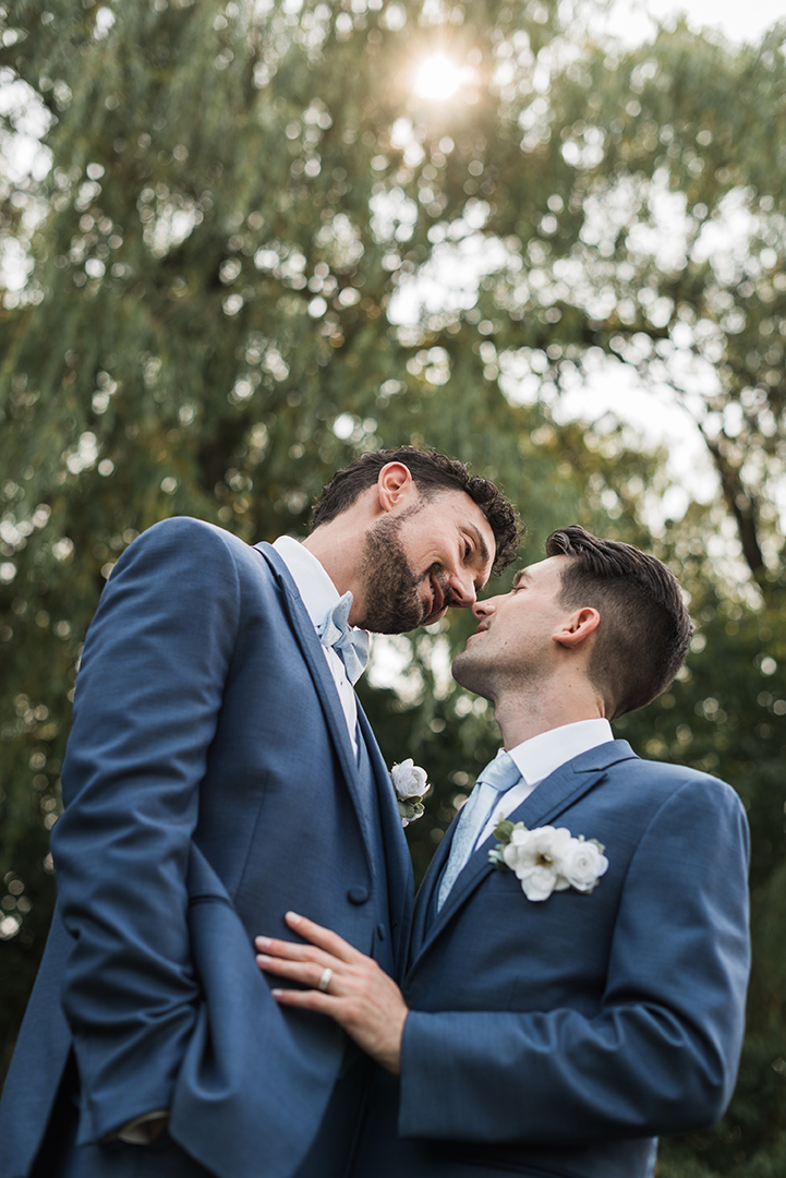 Two grooms - same sex couple - look at each other lovingly in front of trees