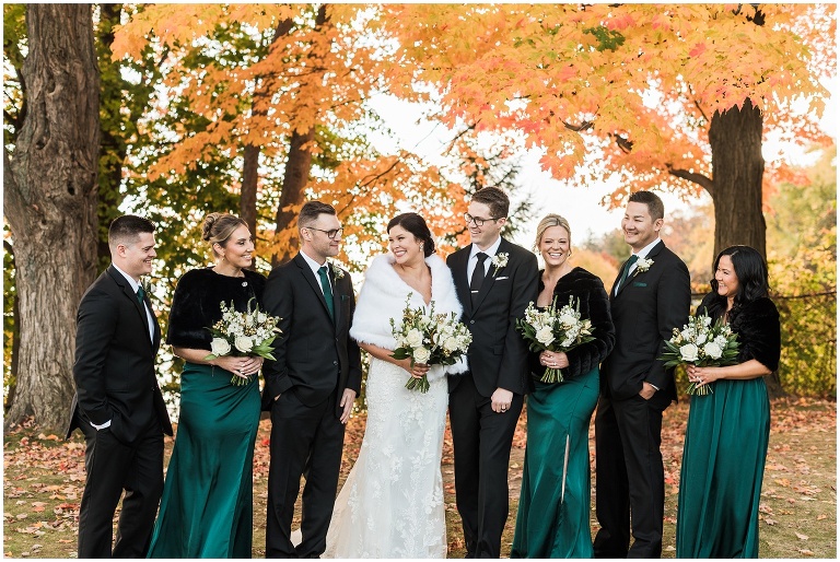 Wedding party dressed in emerald greens, blacks and white flowers standing togethr look at each other in front of orange fall leaves at Paletta Mansion